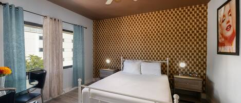 A hotel room with a bed, bedside tables, a desk, a chair, a ceiling fan, and a window with curtains.