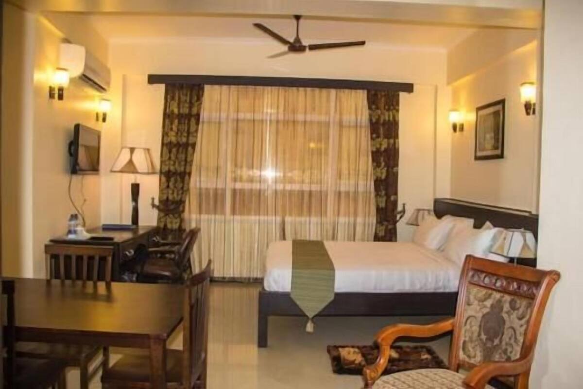 Une chambre d’hôtel équipée d’un lit, d’un bureau, d’une chaise, d’un ventilateur de plafond et d’une fenêtre avec des rideaux.