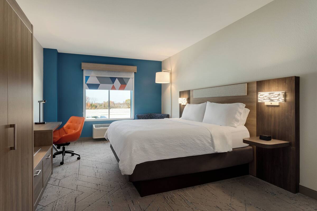 Ein modernes Hotelzimmer mit einem großen Bett, einem Schreibtisch, einem orangefarbenen Sessel und einem Fenster mit geometrischer Aussicht.