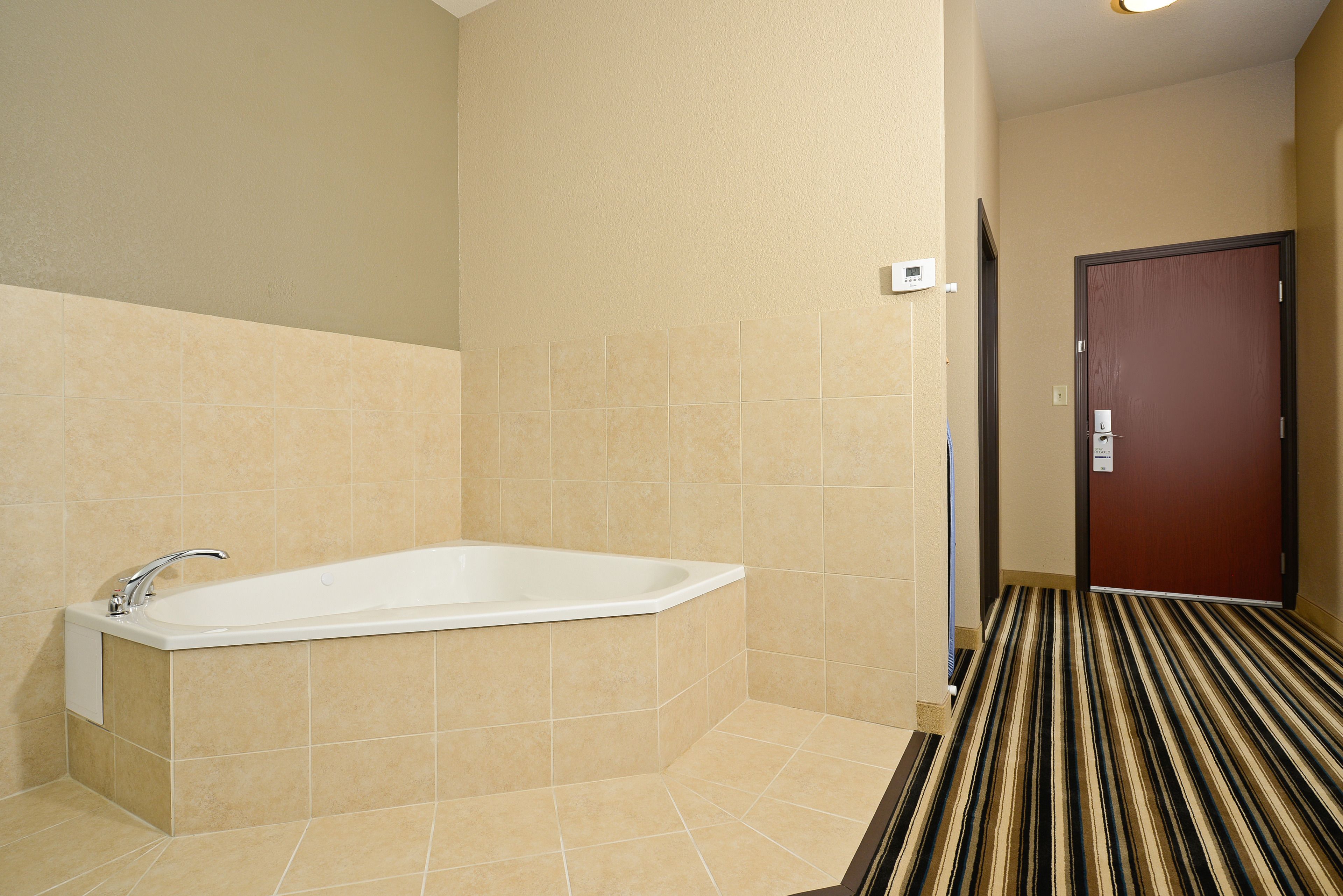 A hotel bathroom with a bathtub, beige tiled walls, and a striped carpeted floor.