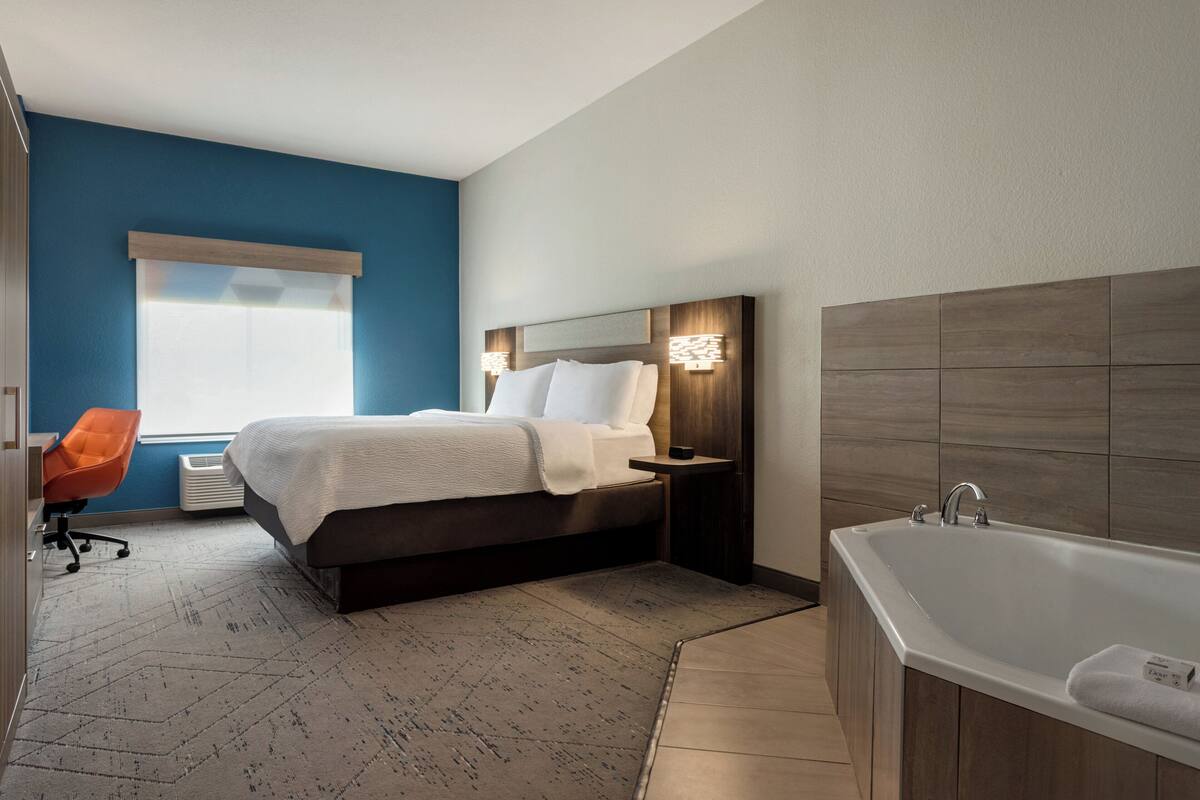 Ein modernes Hotelzimmer mit einem großen Bett, einer Sitzecke mit einem Sessel, einer Badewanne und einem Fenster mit Jalousien.