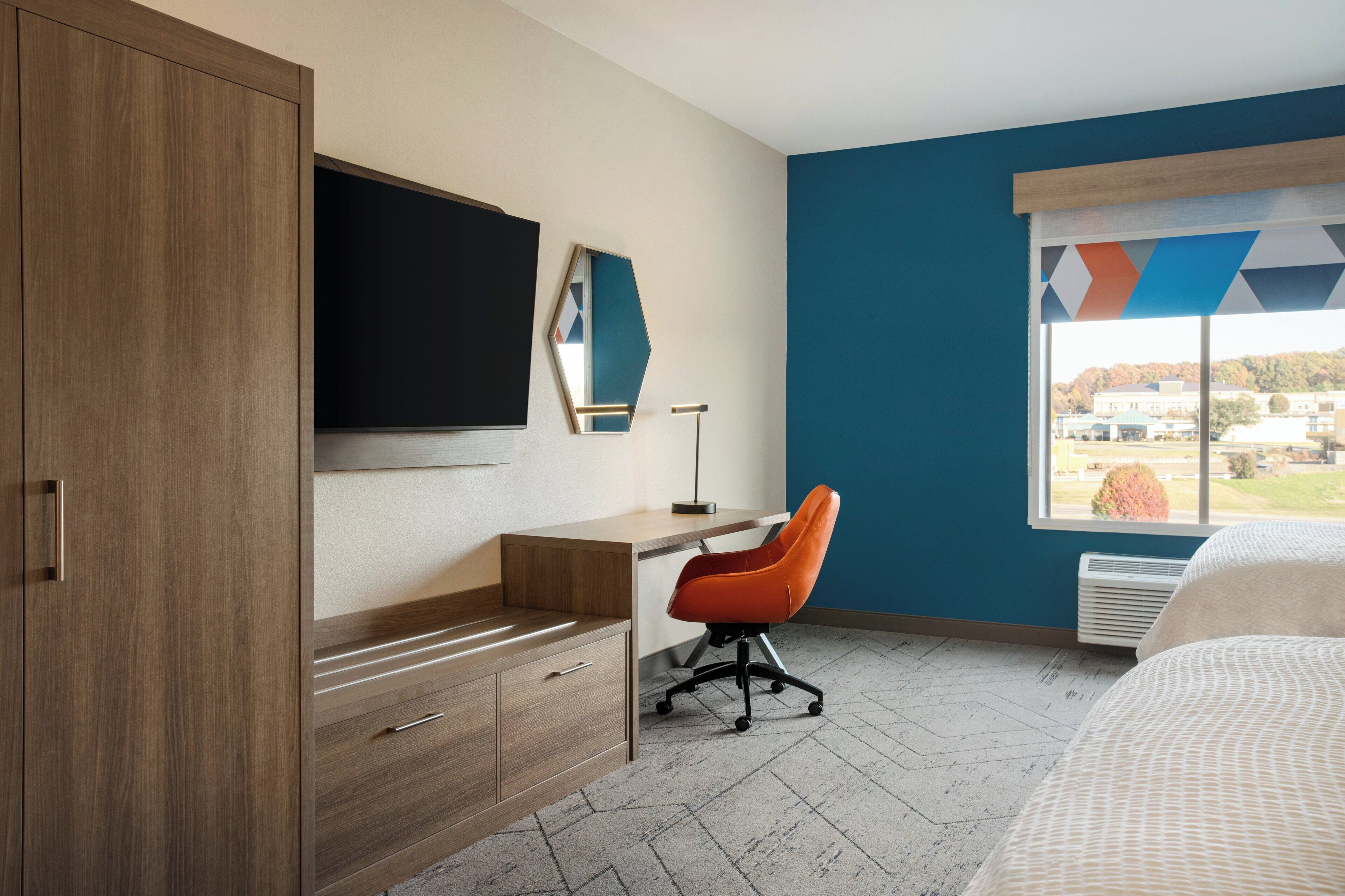 A modern hotel room with a wooden wardrobe, a desk with a chair, a flat-screen TV, and a large window with a geometric view.