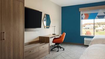 A modern hotel room with a wooden wardrobe, a desk with a chair, a flat-screen TV, and a large window with a geometric view.