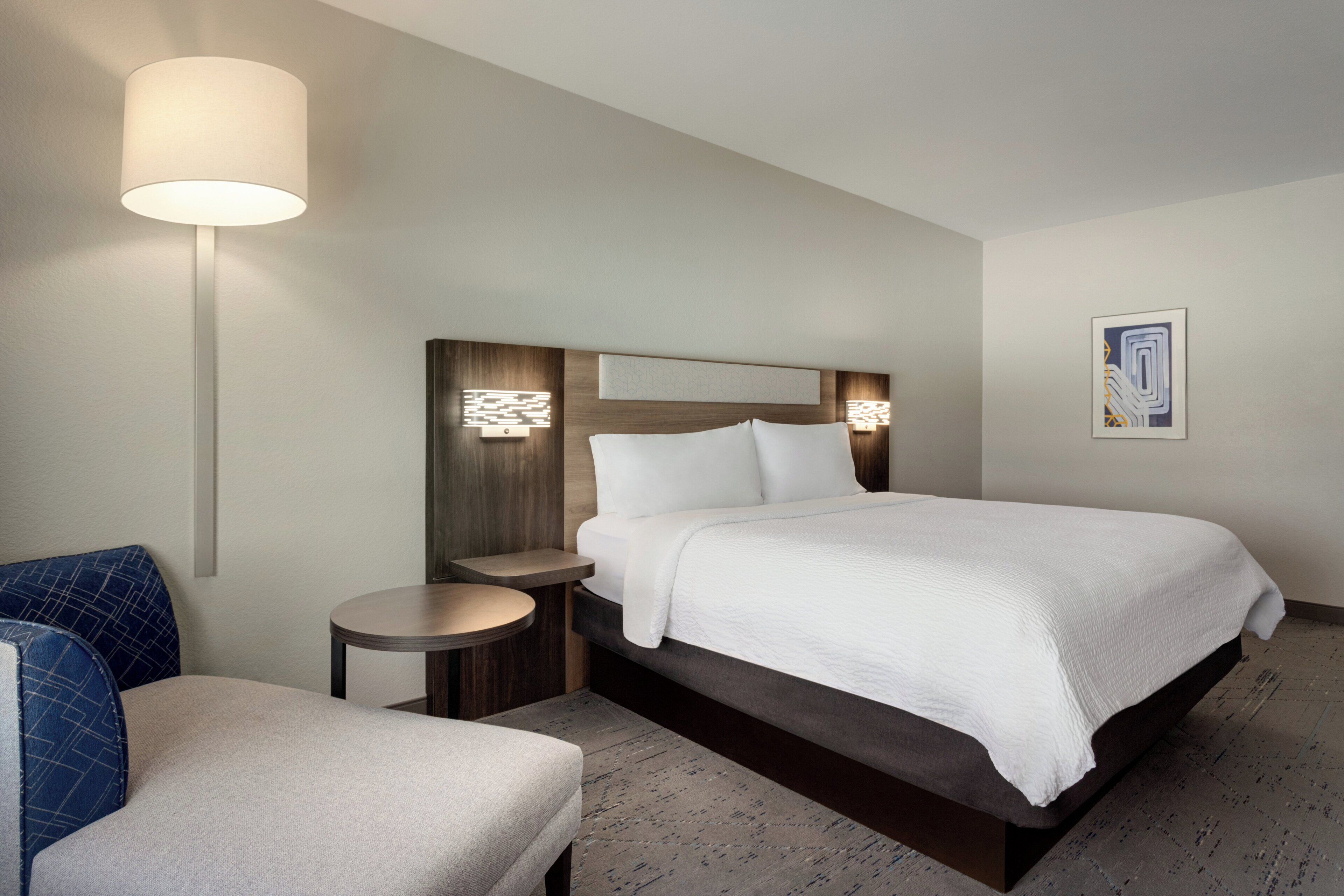 Una habitación de hotel con cama, mesitas de noche, lámpara y una silla.