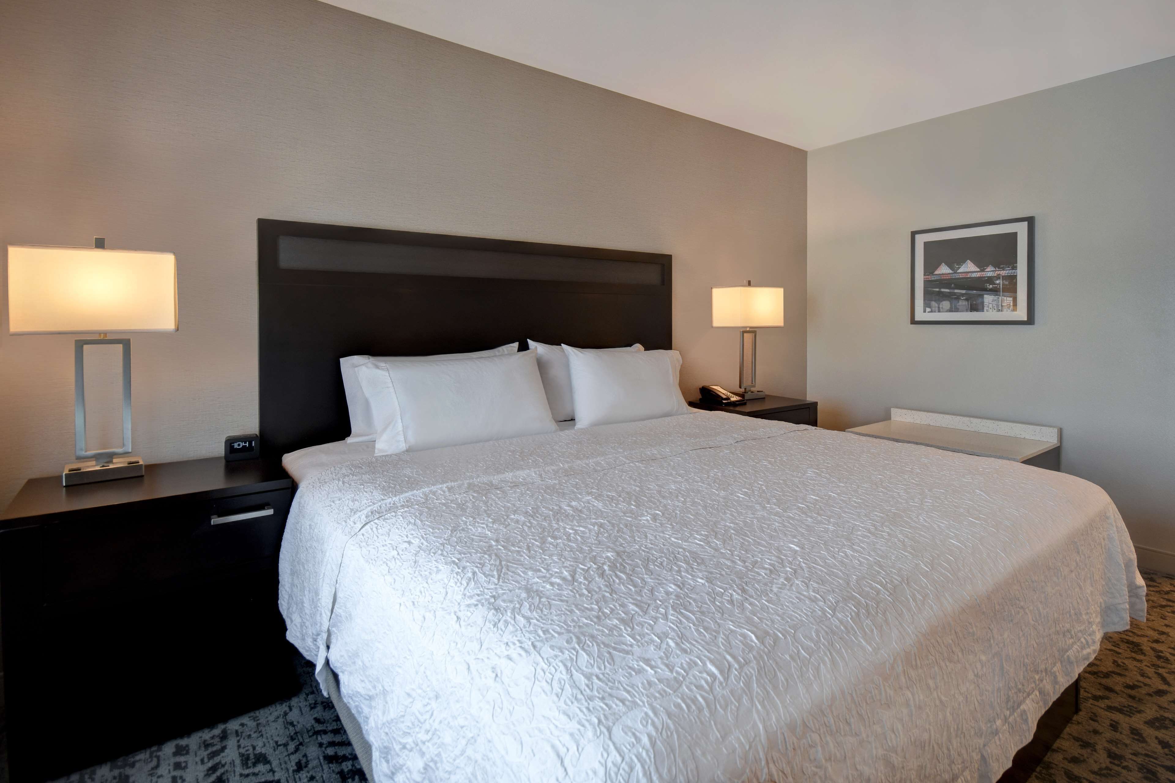 A hotel room with a large bed, two bedside tables with lamps, a nightstand, and a framed picture on the wall.