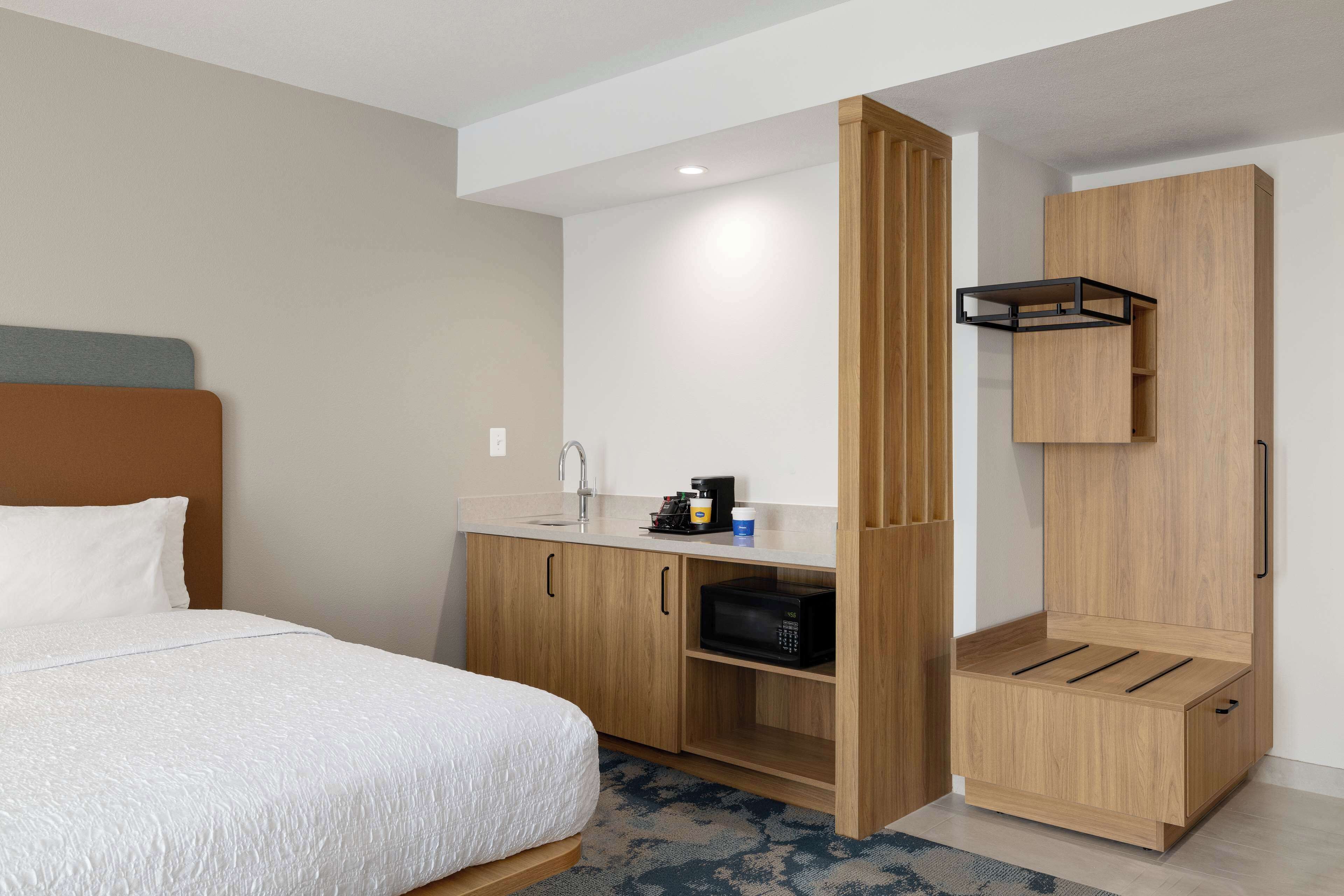 A modern hotel room with a bed, a wooden wardrobe, a built-in microwave, and a small kitchenette area.