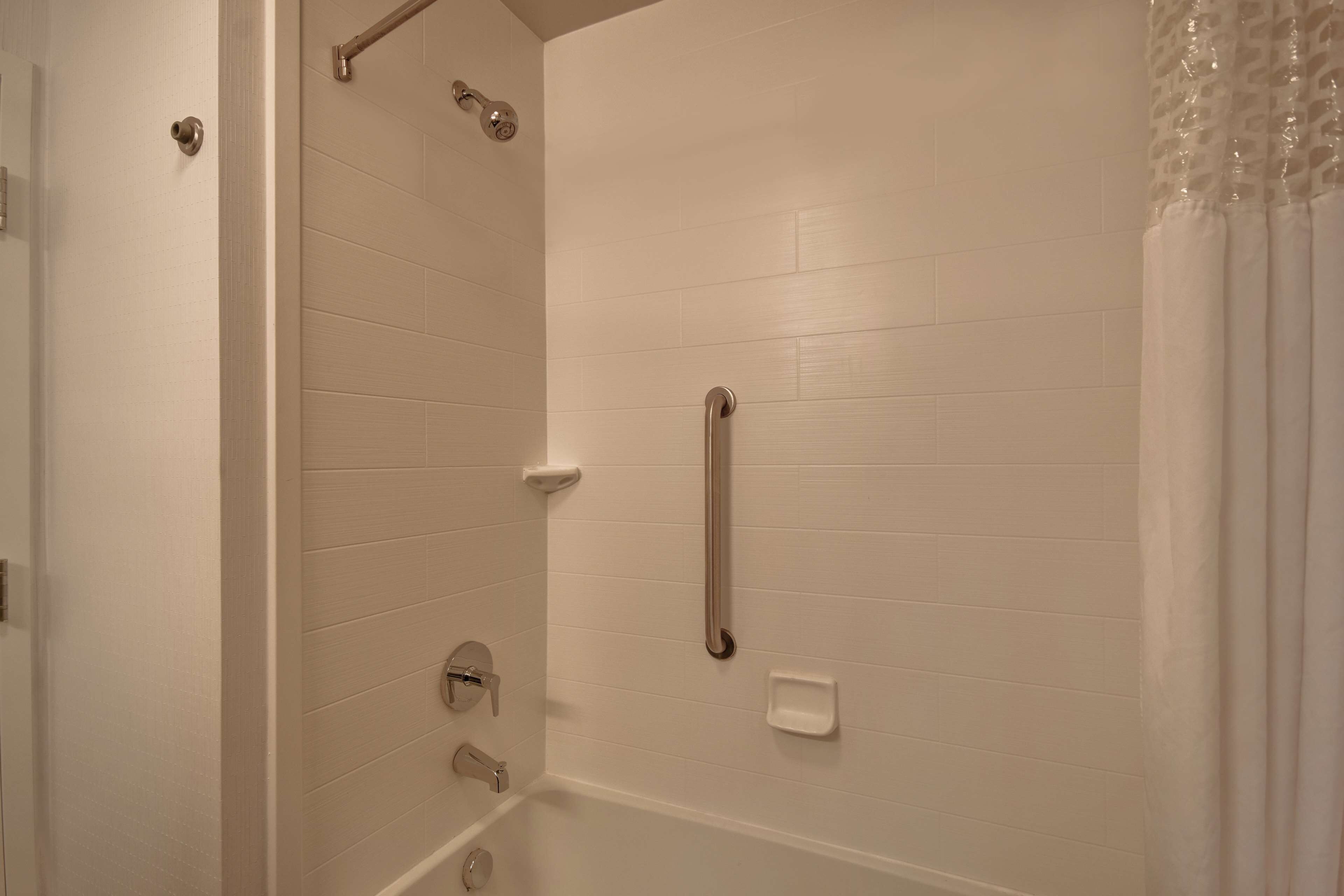 A bathroom with white tiled walls, a shower with a handrail, and a toilet with a grab bar.