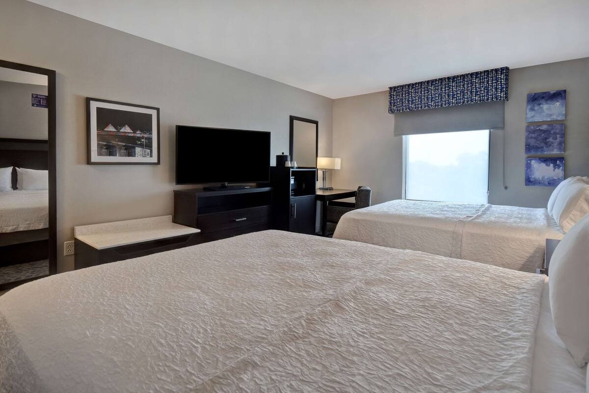 A hotel room with two beds, a TV, a desk, and a window with a view.