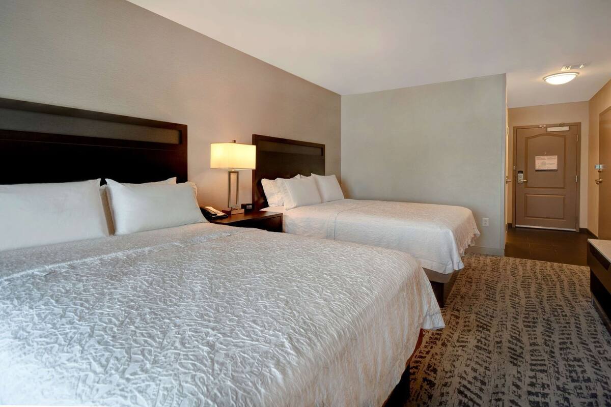 A hotel room with two beds, a nightstand, a lamp, and a door.