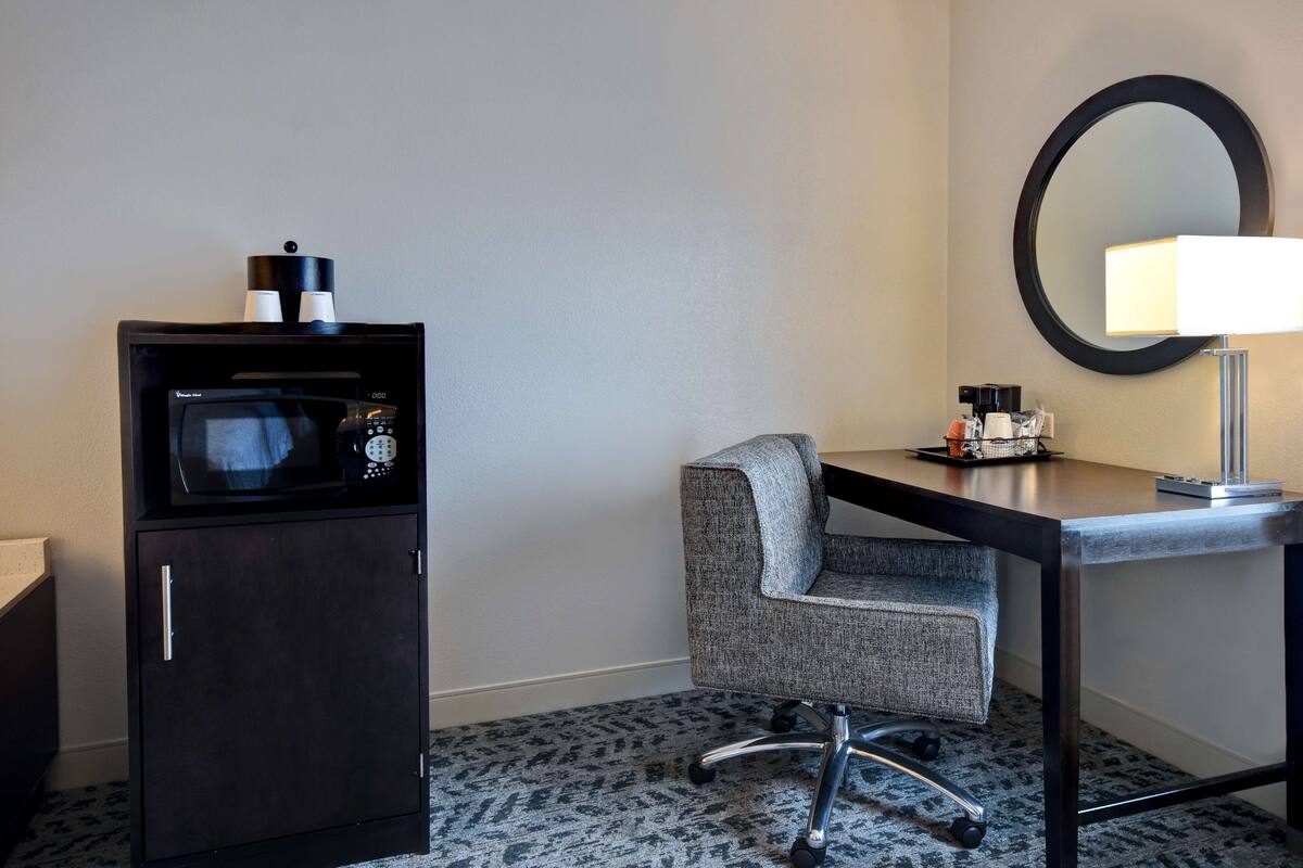 A compact hotel room with a kitchenette, a desk with a lamp, a chair, and a large mirror.