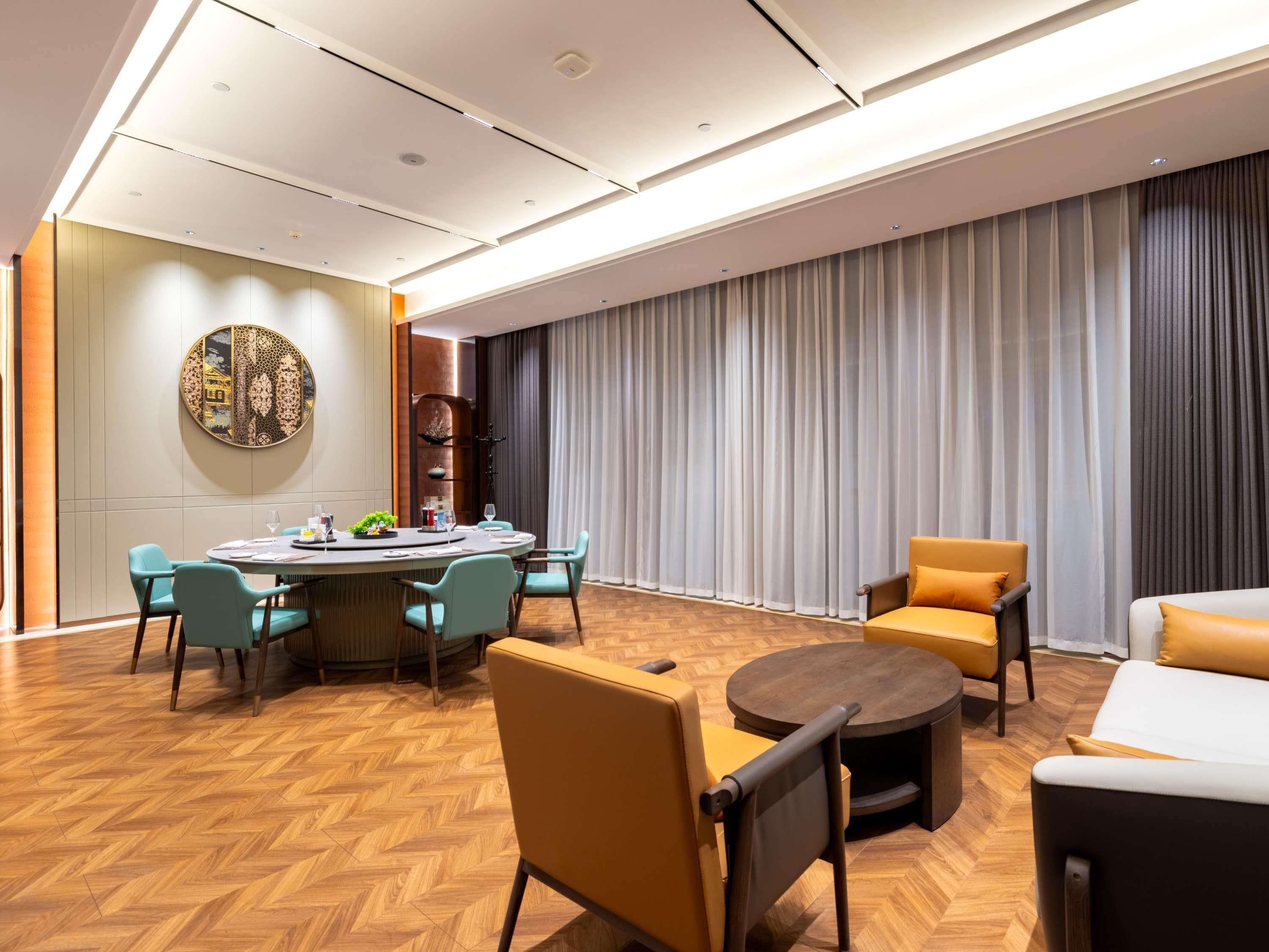 A modern hotel room with a round dining table, teal chairs, a sofa, and a large window with curtains.