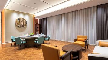 A modern hotel room with a round dining table, teal chairs, a sofa, and a large window with curtains.