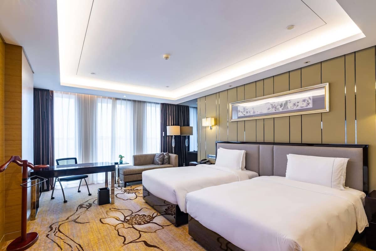 A hotel room with two beds, a desk, a chair, a lamp, and a large window with curtains.