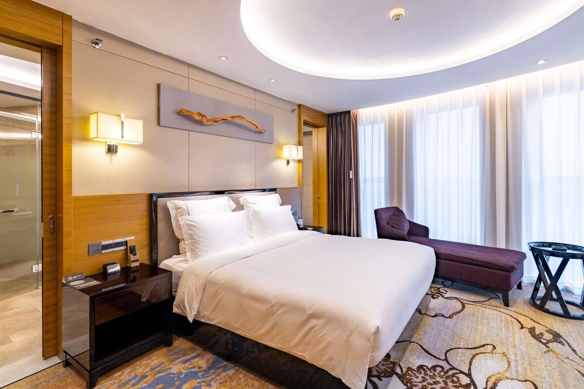 A hotel room with a bed, bedside table, lamp, chair, and a view of the outdoors.