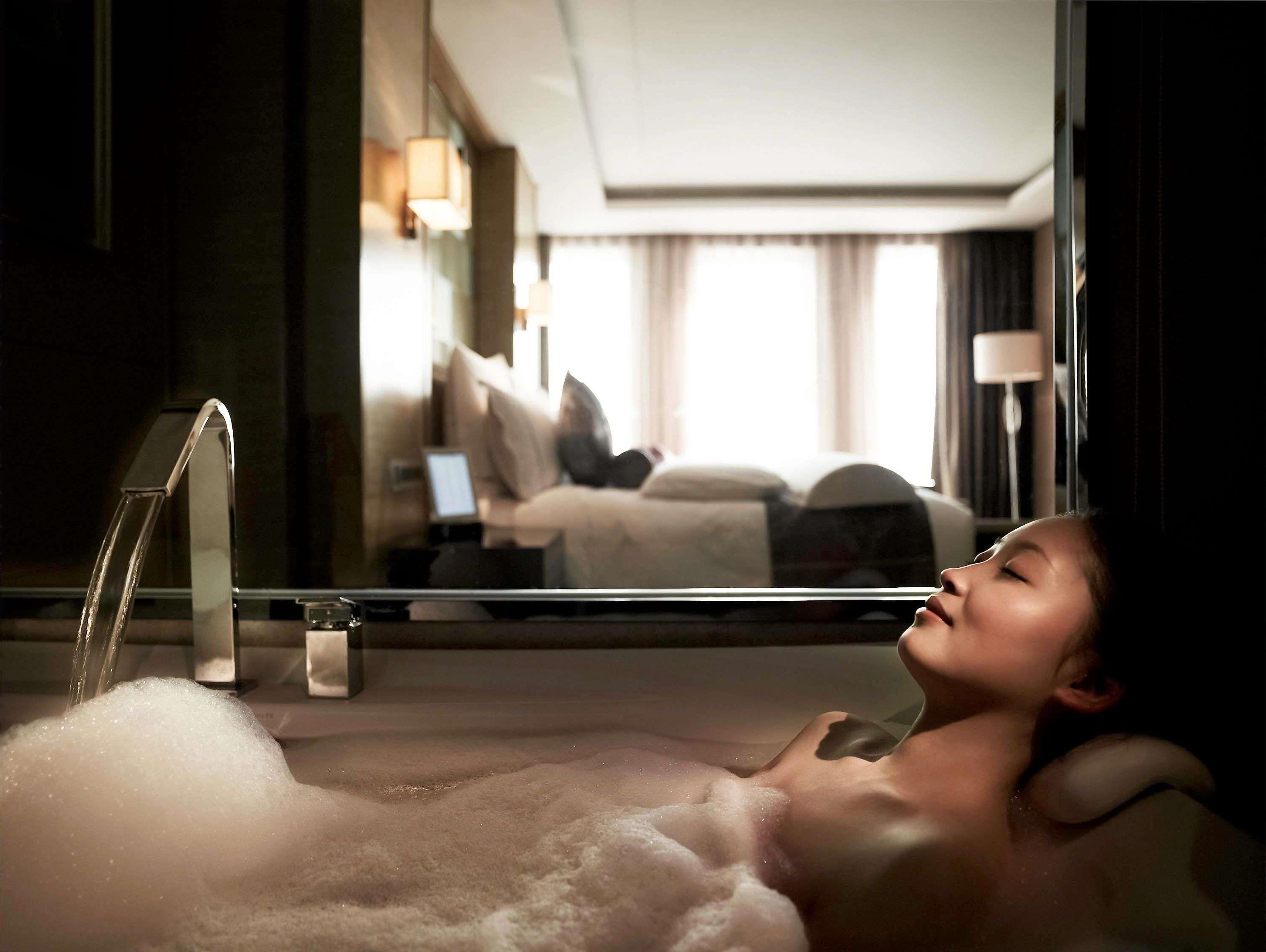 Una persona relajándose en una bañera con vista a una habitación de hotel, reflejada en un espejo.