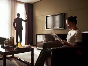 Ein Wohnzimmer mit einer Couch, einem Couchtisch mit einer Auflage und verschiedenen Gegenständen, einem Fernseher und einer Person, die am Fenster steht.