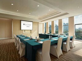 Un salón de conferencias con una larga mesa, sillas, una pantalla de proyector y vistas a edificios a través de amplias ventanas.