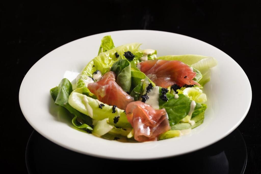 A white plate with a salad of mixed greens, prosciutto, and black olives, served on a black background.