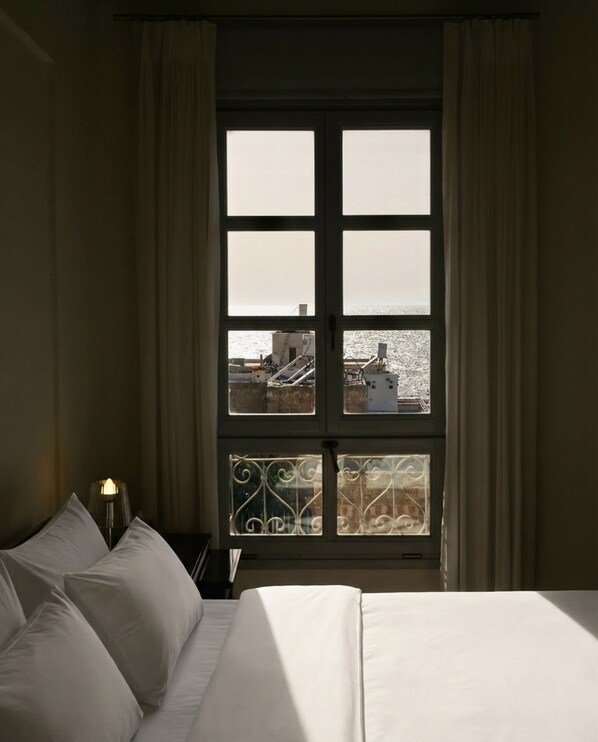 A hotel room with a bed, a nightstand with a lamp, and a window with a view of buildings and water. - The Efendi Hotel (Acre)