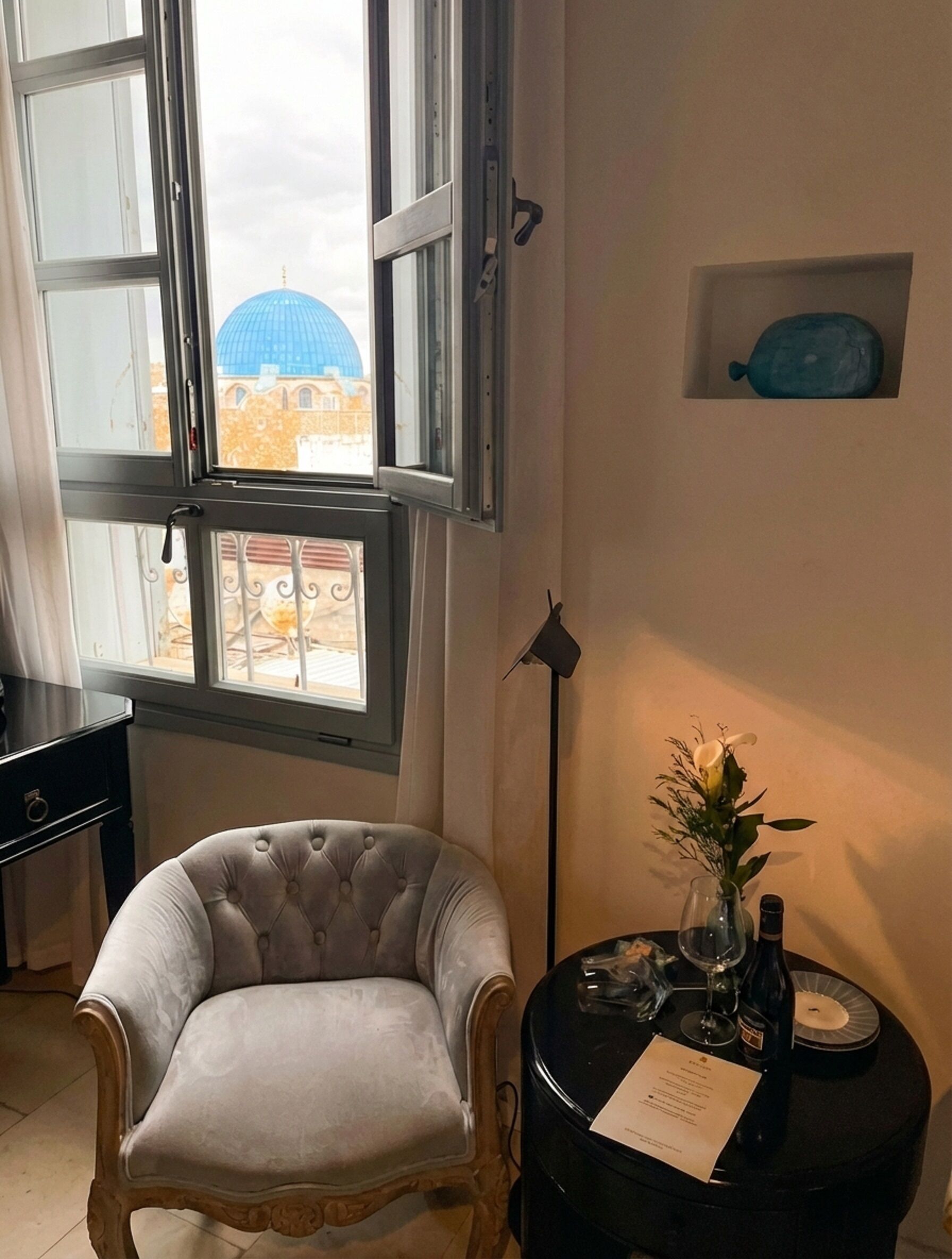 A room with a large window, a chair, a small table with a vase and a bottle, and a wall-mounted shelf with a blue object.