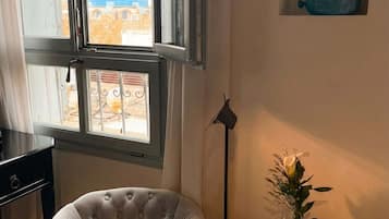 A room with a large window, a chair, a small table with a vase and a bottle, and a wall-mounted shelf with a blue object.