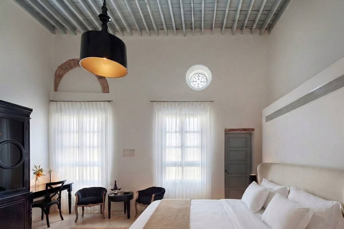 A hotel room with a bed, two chairs, a small table, a desk, a lamp, and a ceiling light.
