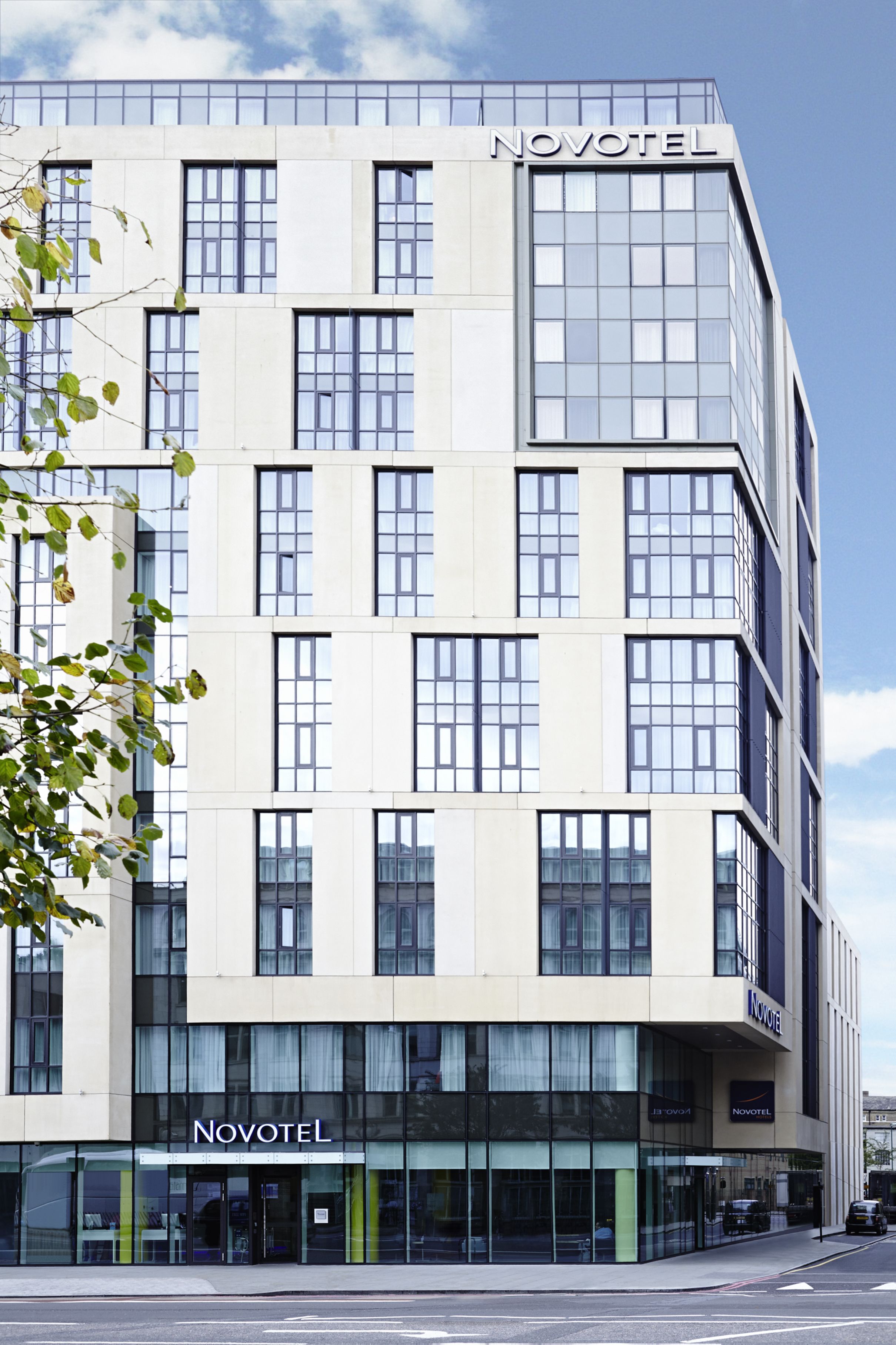 A modern Novotel hotel building with a prominent glass facade, multiple floors, and a clear sky in the background.