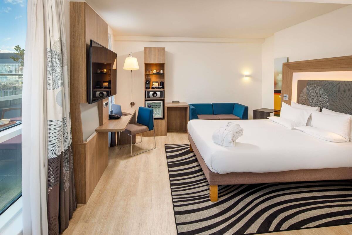 A hotel room with a bed, a desk, a chair, a TV, and a minibar.
