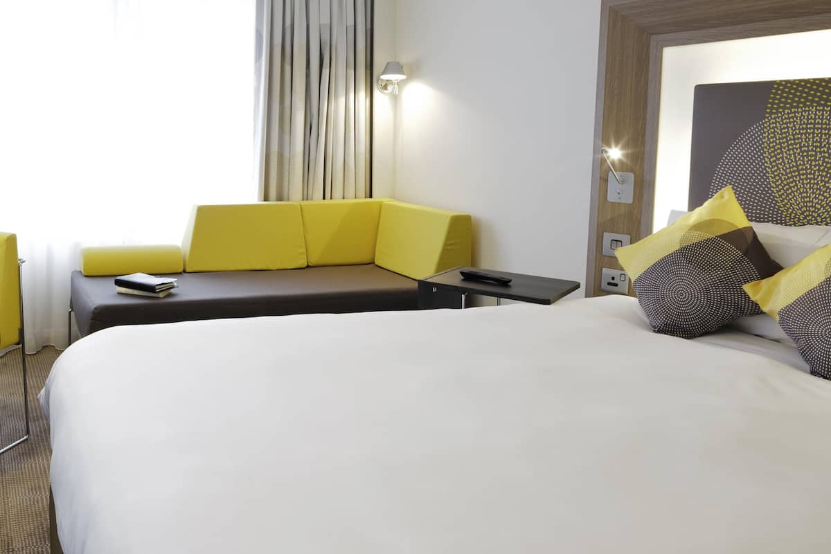 A hotel room with a large bed, a sofa, a bedside table, a lamp, and a window with curtains.