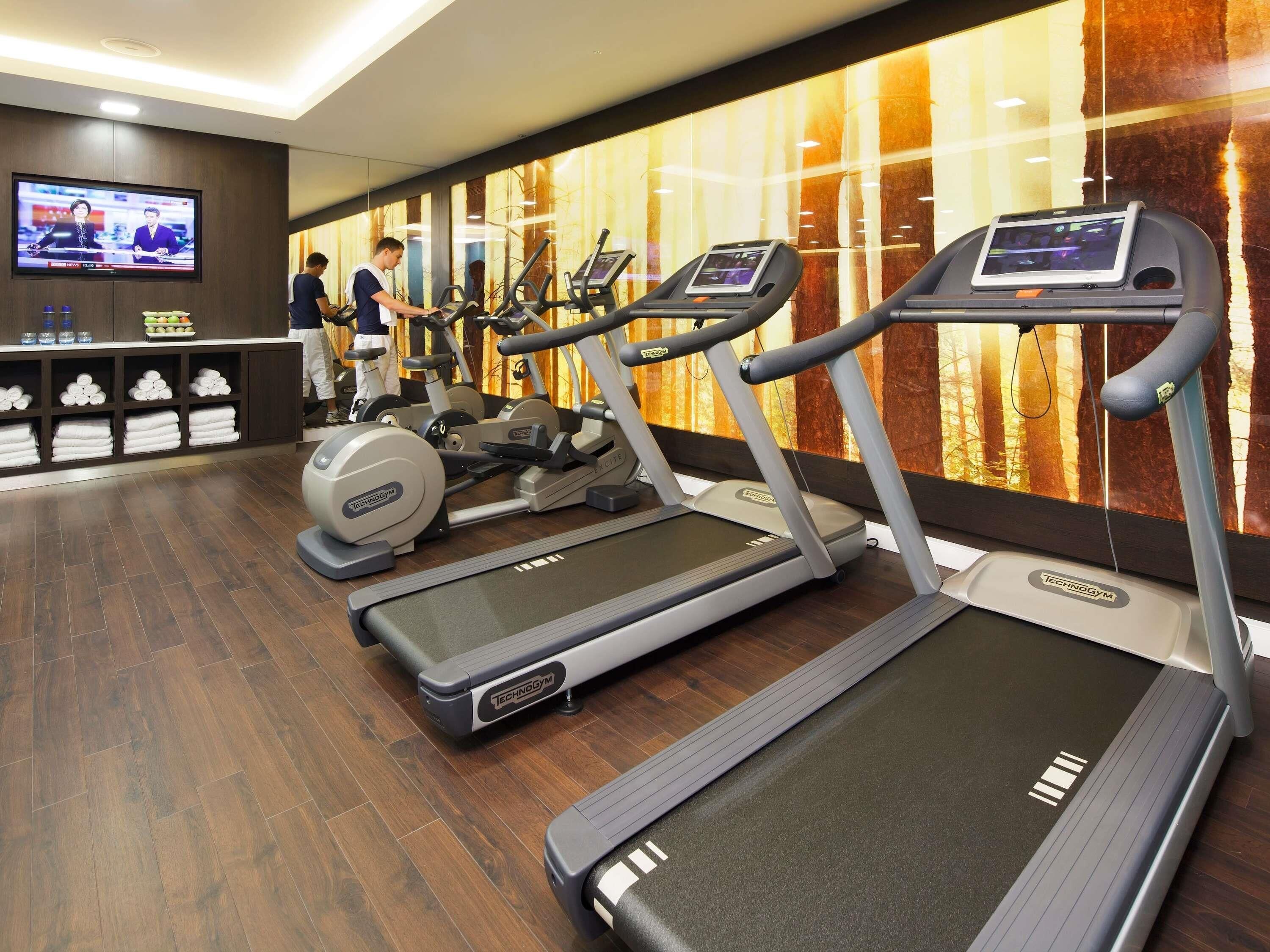 Ein Fitnessstudio mit Laufbändern, Crosstrainern und einem Fernseher, der eine Nachrichtensendung zeigt.