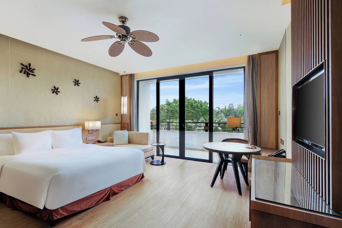 A hotel room with a large bed, a ceiling fan, a TV, a balcony with a view of trees, and a small table.