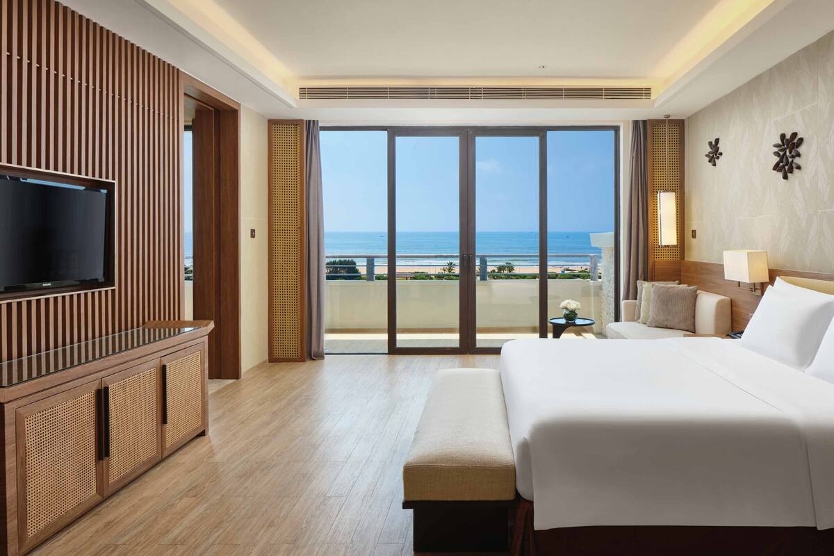 A hotel room with a large bed, a TV, and a balcony with a view of the beach.