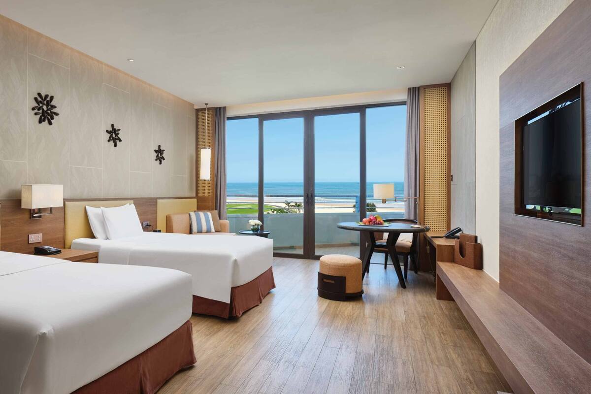 A hotel room with two beds, a dining area with a table and chairs, a TV, and a large window with a view of the beach.