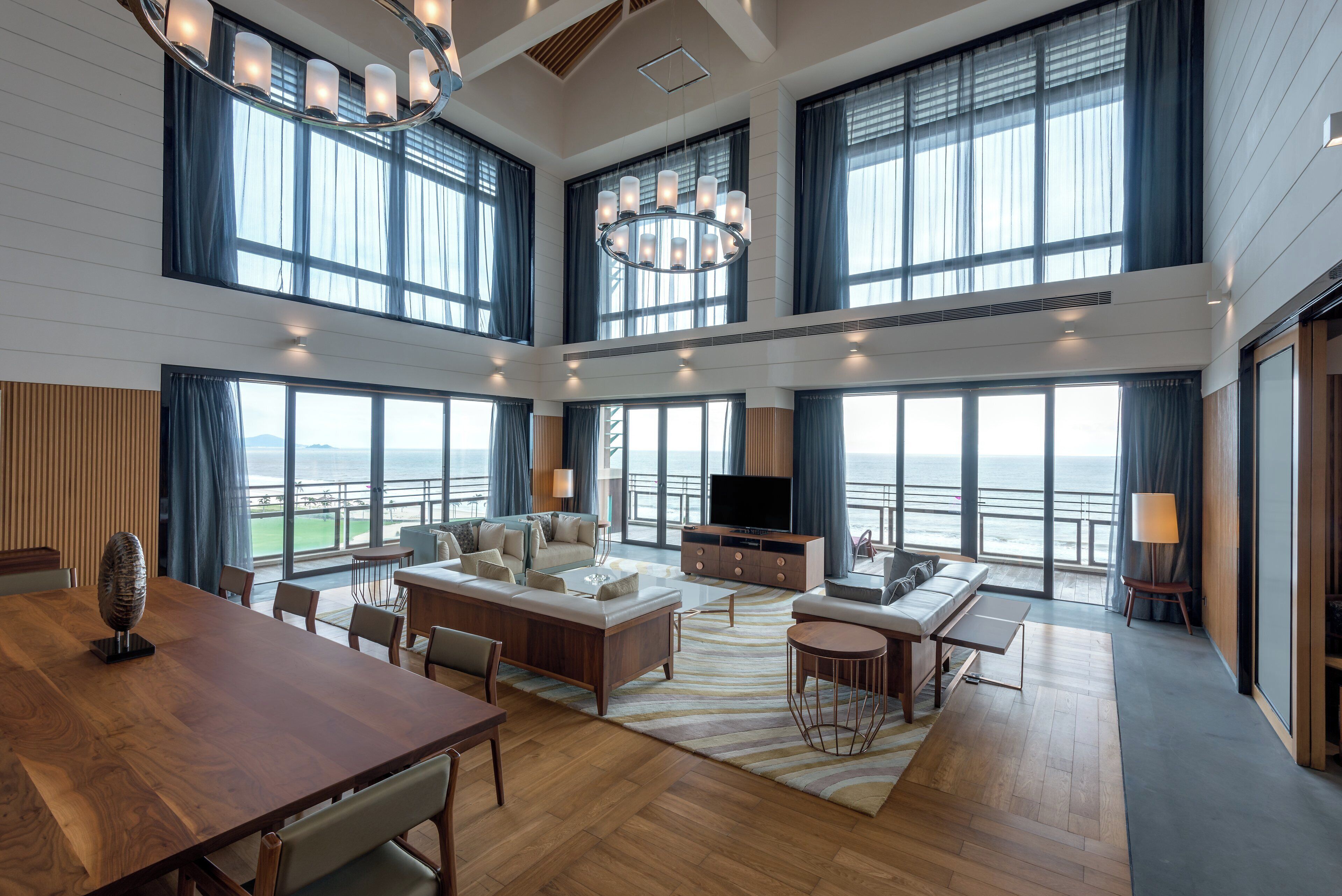 A spacious living area with a large window overlooking the sea, a modern chandelier, and a television on a wooden cabinet.