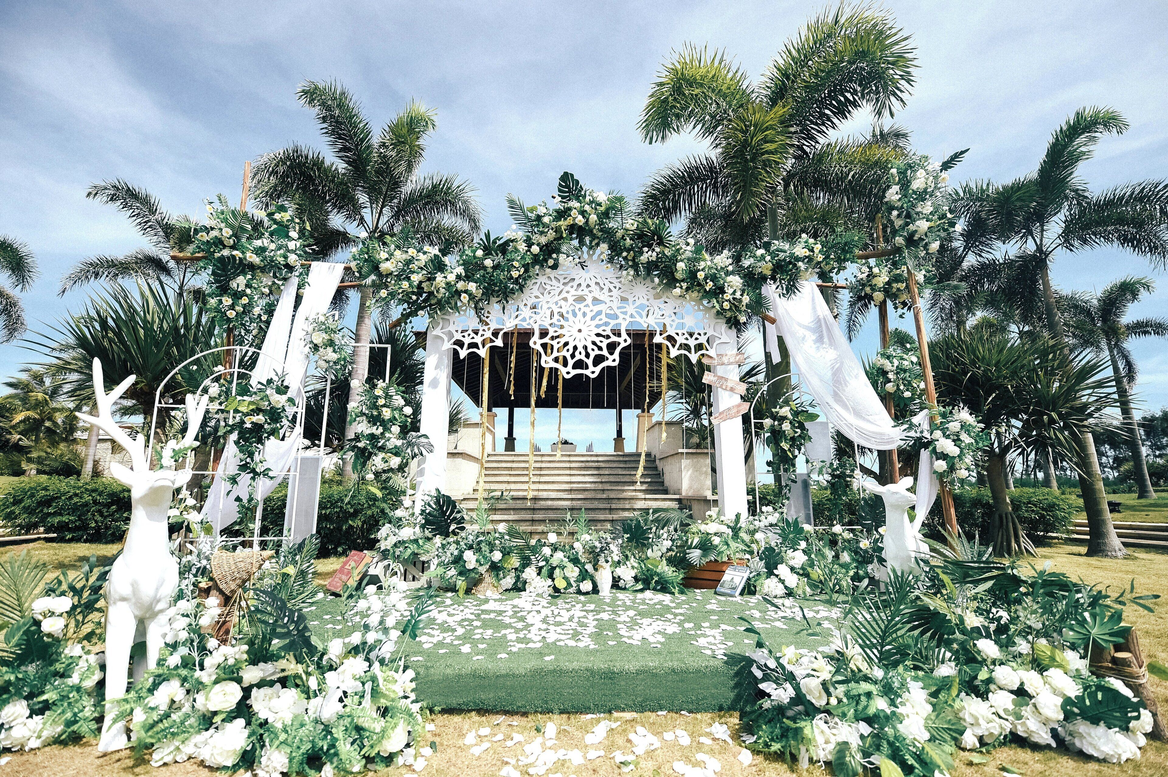 Une arche de cérémonie nupciale décorée de fleurs blanches et de verdure, entourée de palmiers et d’une pelouse.