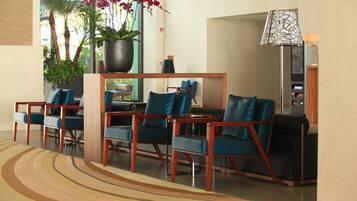 A lobby with a seating area featuring wooden chairs and a table, a potted plant with flowers, and a lamp.