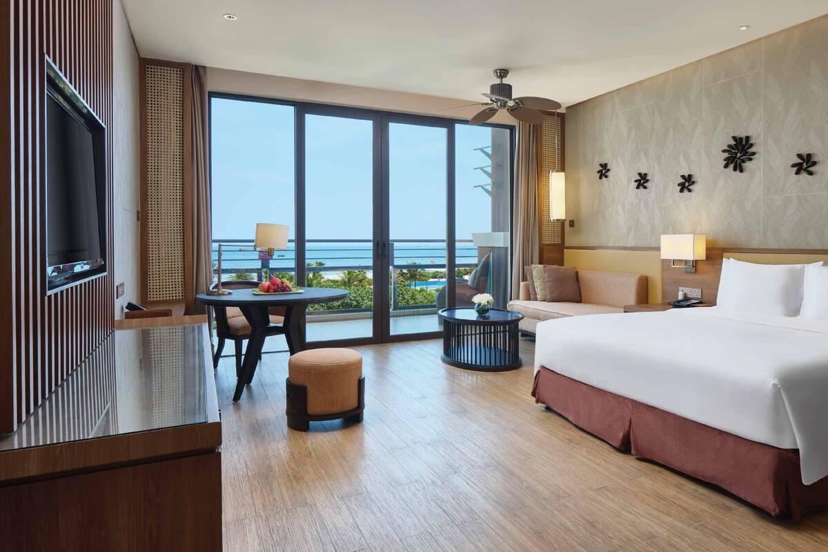 A hotel room with a large bed, a desk with a TV, a dining table with fruit, and a view of the ocean.
