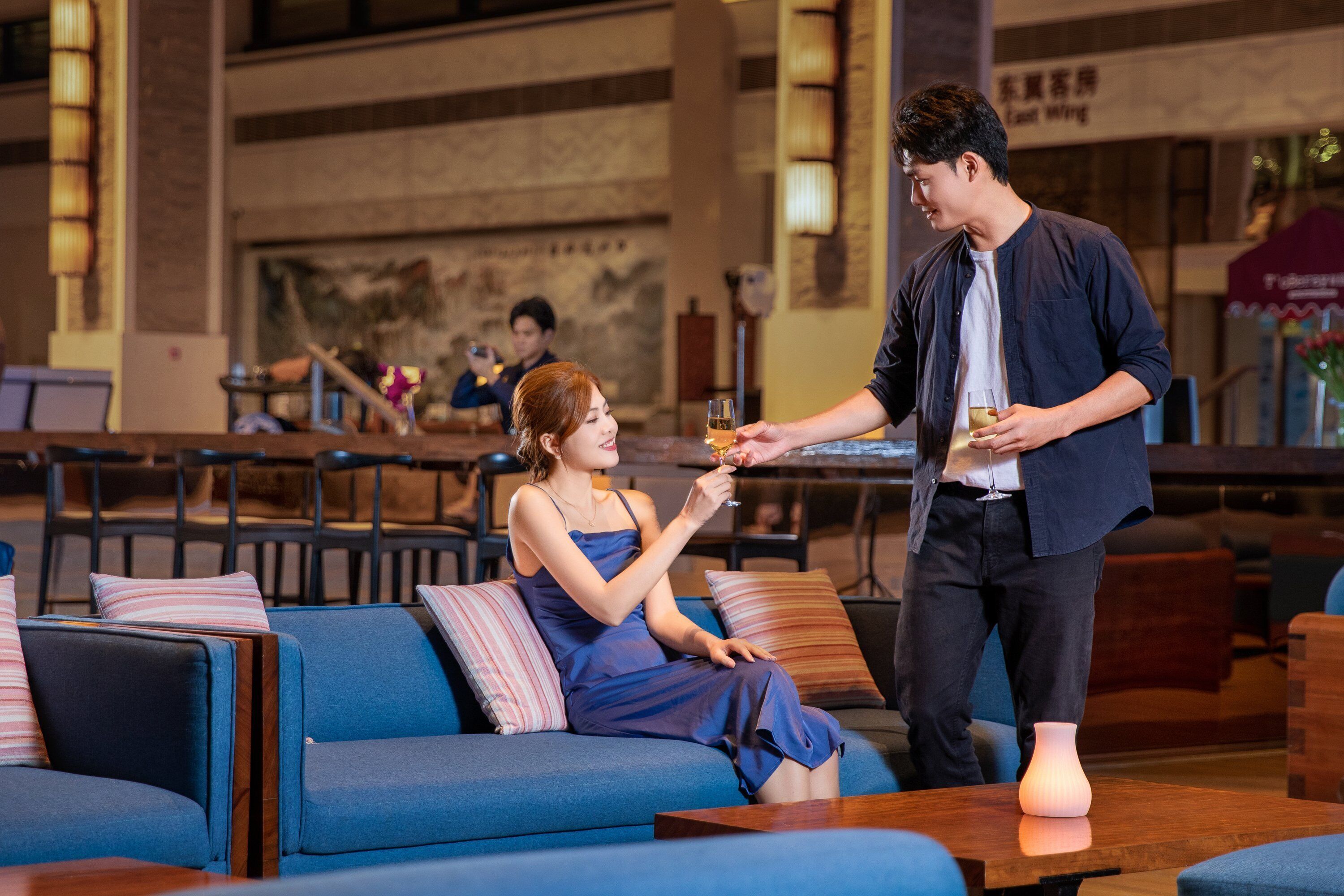A person serving drinks to another person seated on a sofa in a restaurant setting.