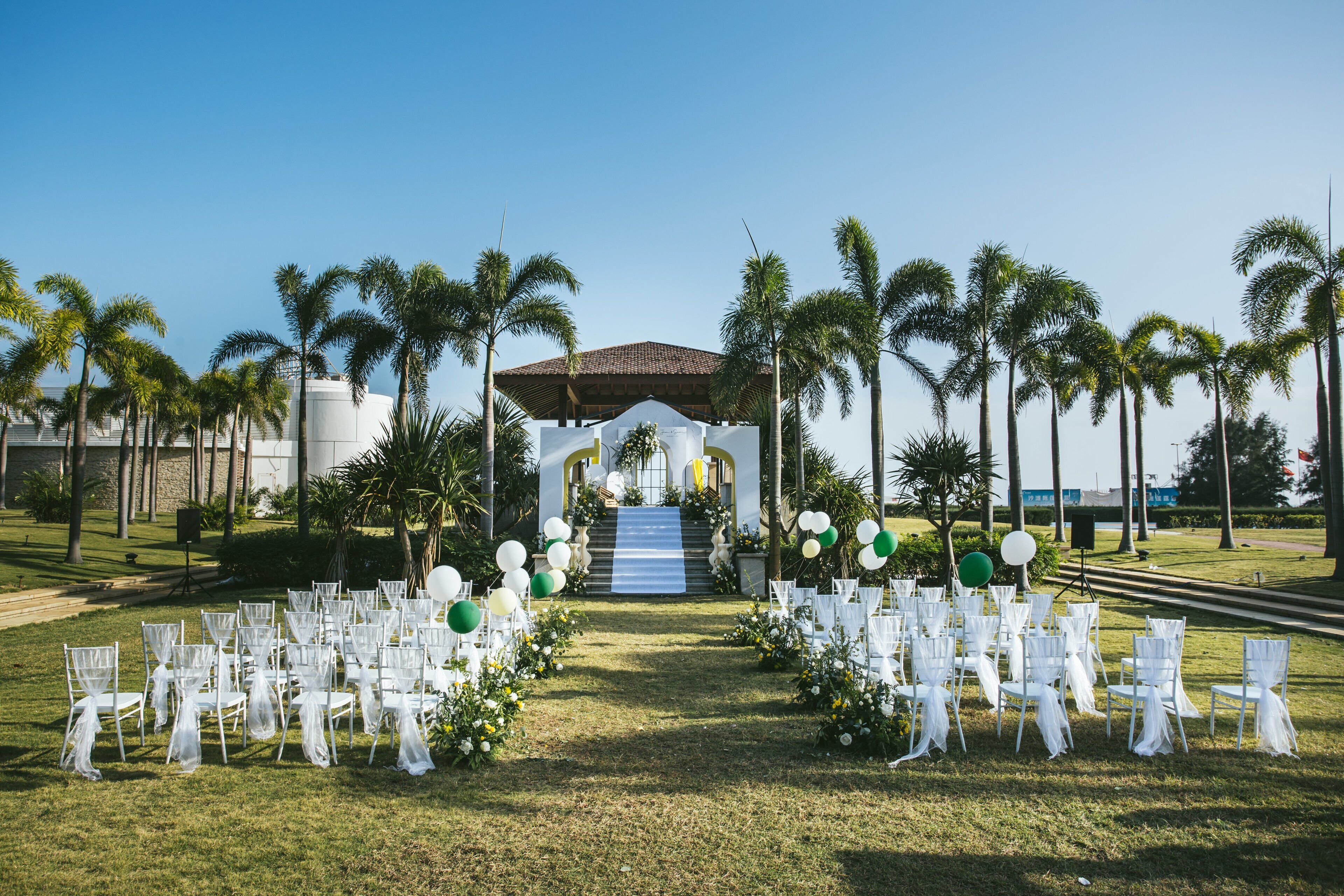 An outdoor wedding venue with white chairs arranged in rows, a staircase leading to a covered area, and palm trees in the background.