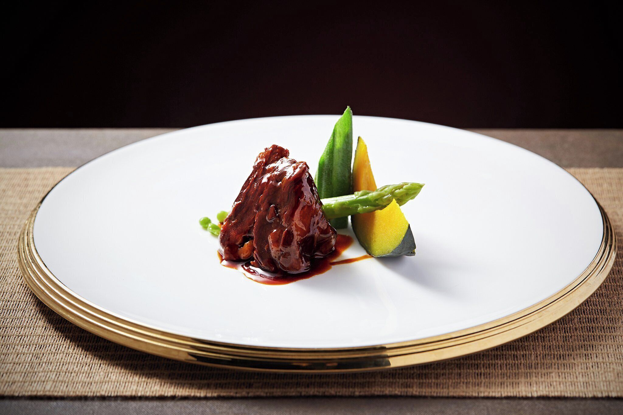 A white plate with a piece of meat, asparagus, and a slice of mango on a tablecloth.