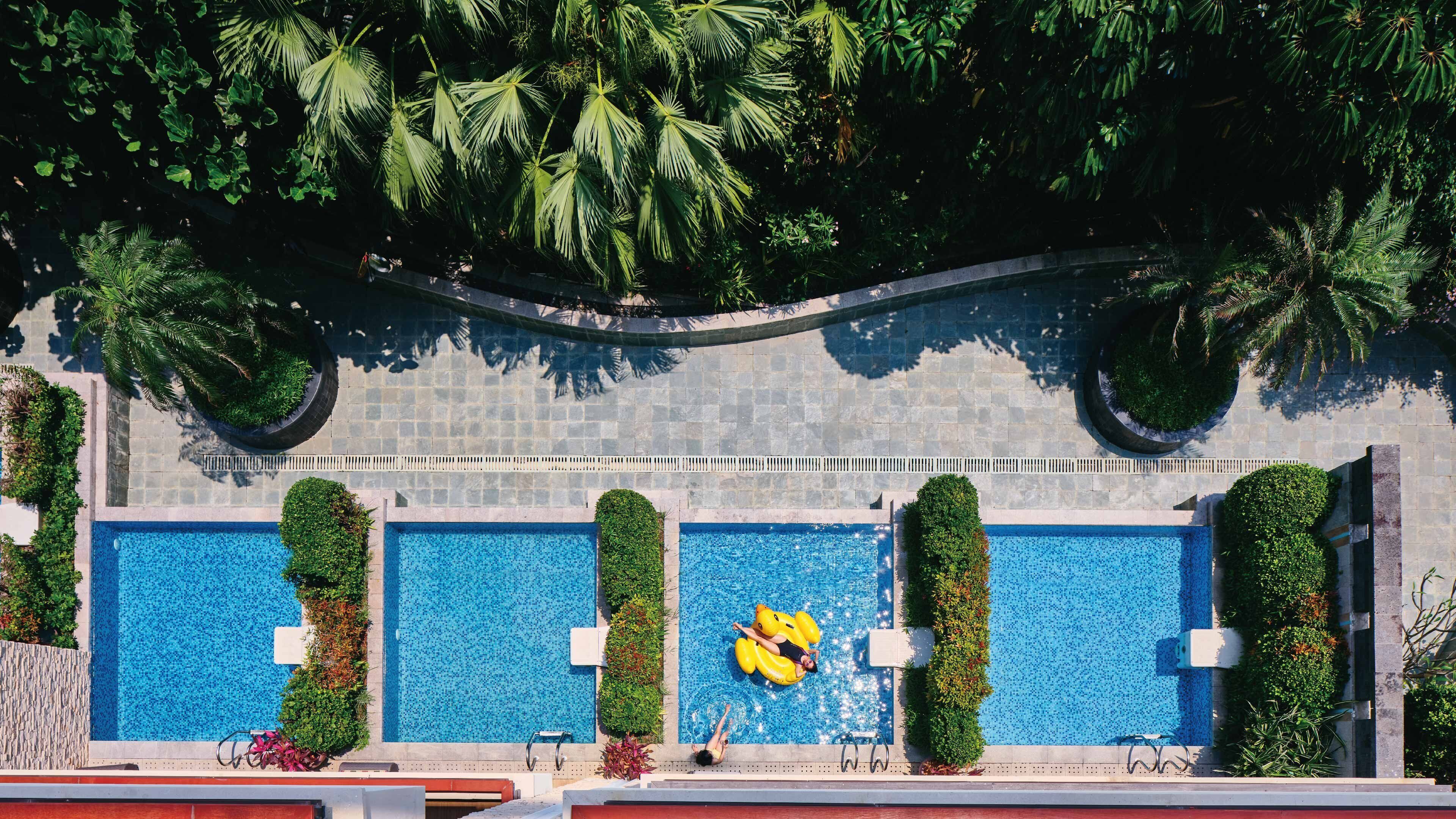 A rooftop pool area with three swimming pools, surrounded by lush greenery and palm trees.