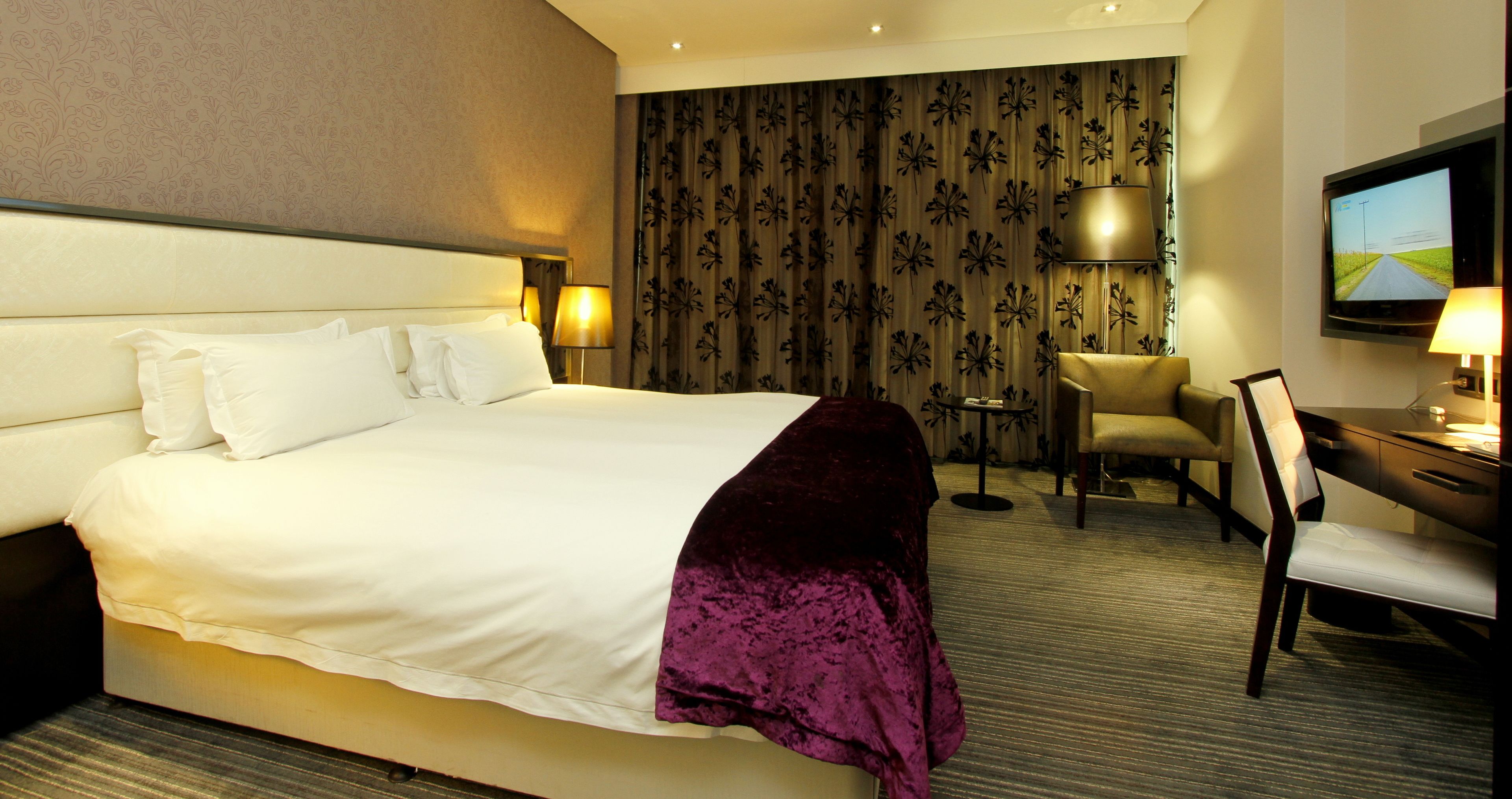 A hotel room with a large bed, a desk with a chair, a TV, and a small table with a lamp.