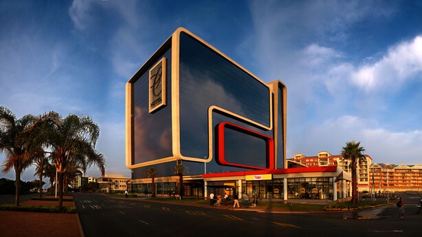Un hotel moderno con una fachada geométrica grande y llamativa, una entrada central y un estacionamiento con palmeras.