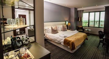 Habitación de hotel con una cama grande, zona de estar con una silla y una mesa, un espejo y vistas a la ciudad.