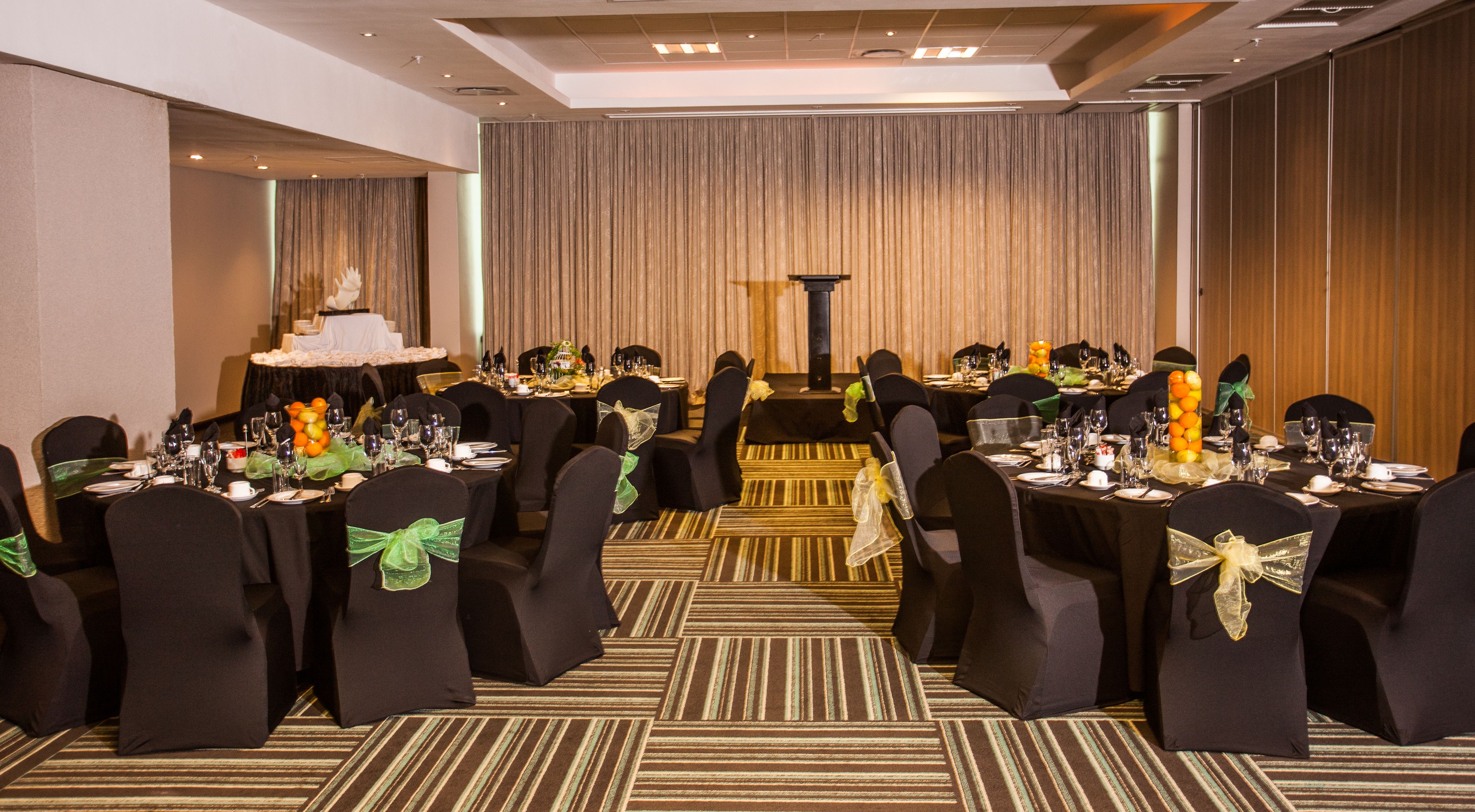 Une salle de banquet avec de nombreuses tables rondes, des chaises noires ornées de rubans verts et un tapis à rayures.