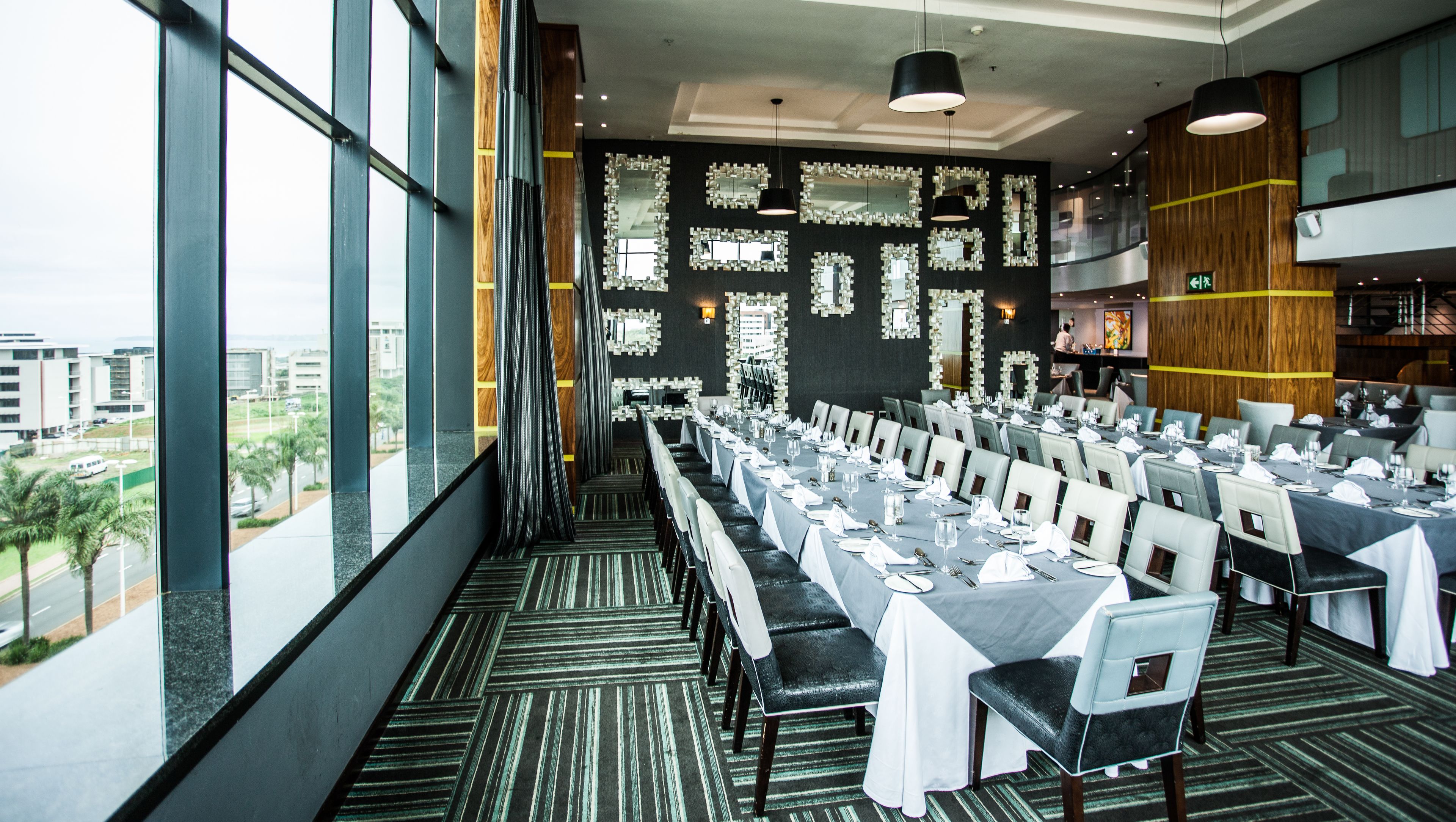 A restaurant with a long table set for a meal, large windows, and a view of the city.