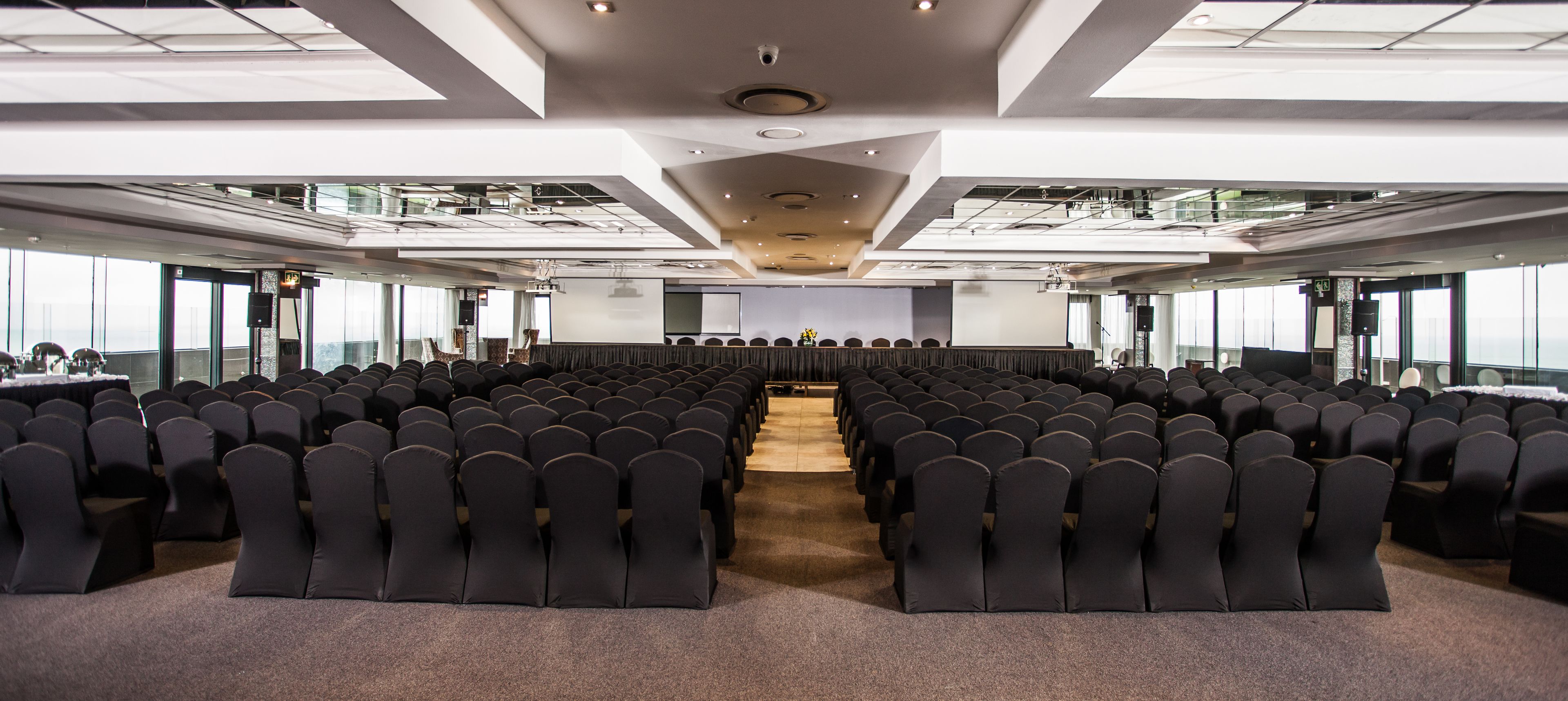 A conference room with rows of chairs, a stage with a screen, and large windows offering a view of the city.