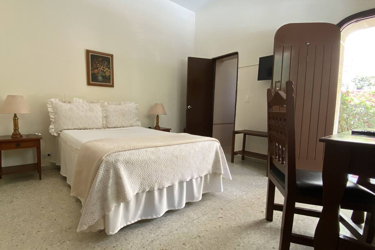 A bedroom with a bed, two bedside tables with lamps, a wooden wardrobe, and a small table with a chair.