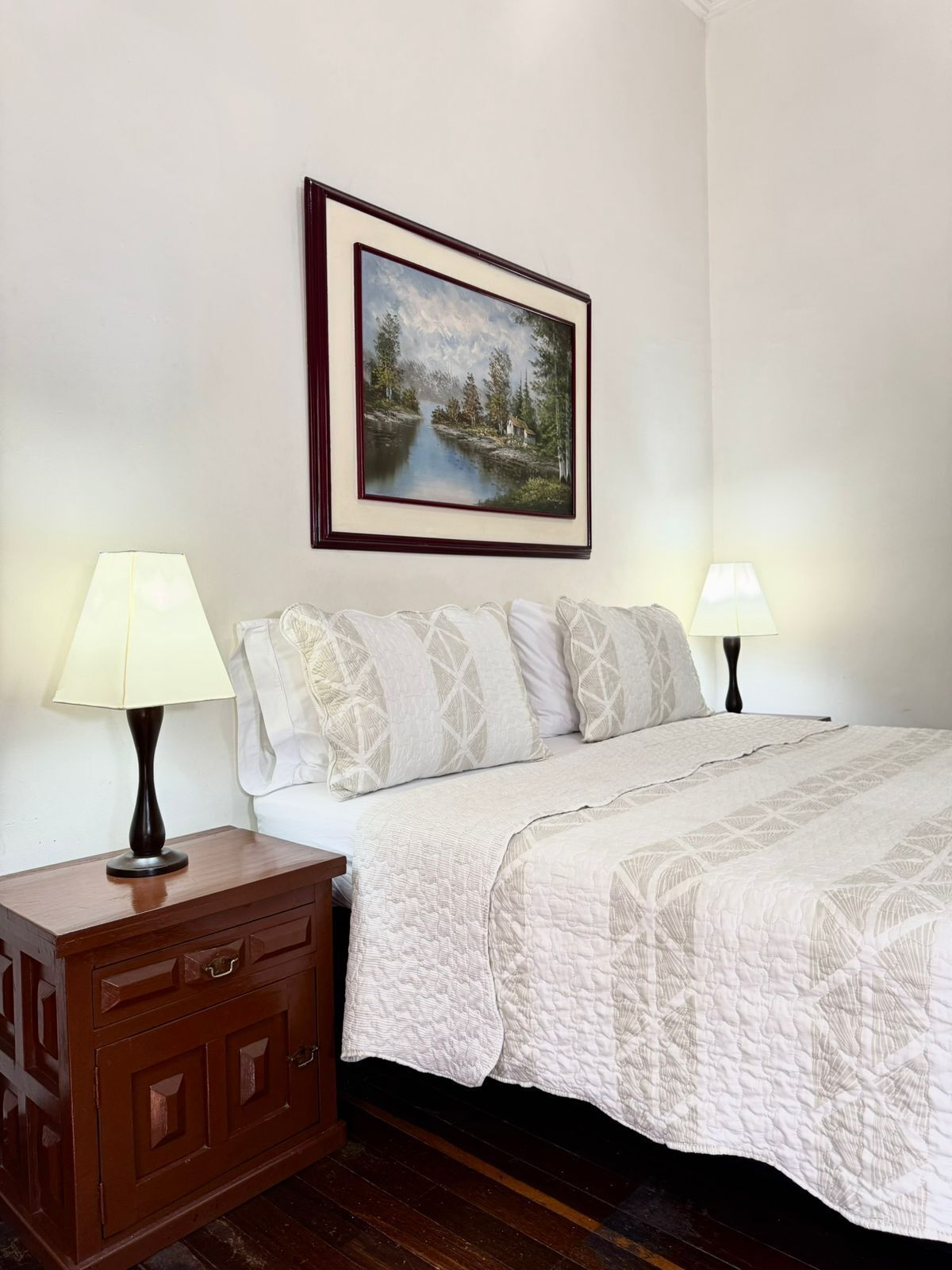 A neatly arranged bedroom with a bed, two bedside lamps, a nightstand, a drawer unit, and a framed landscape painting on the wall.