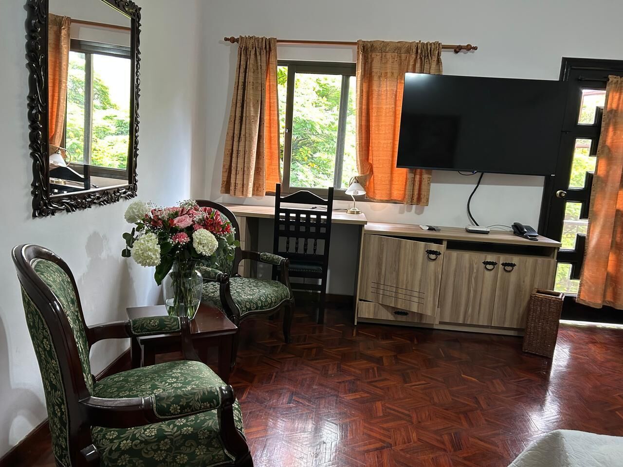 A room with a wooden herringbone floor, a dining table with chairs, a TV mounted on the wall, and a window with curtains.